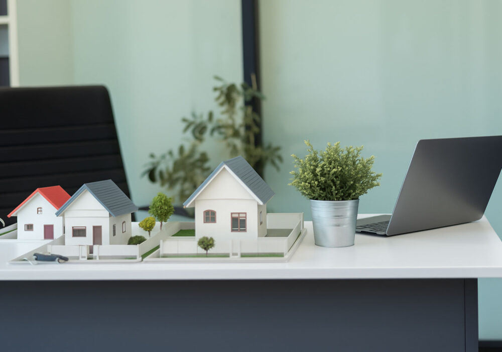 Model houses on a desk with plants and a laptop.